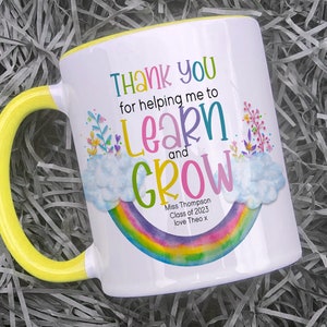 May include: White ceramic mug with a yellow handle. The mug features a rainbow design with clouds and flowers. The text on the mug reads "Thank you for helping me to learn and grow. Miss Thompson Class of 2023 love Theo x".