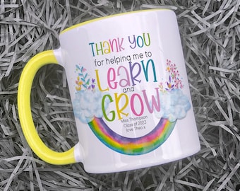 May include: White ceramic mug with a yellow handle. The mug has a rainbow design with clouds and flowers. The text on the mug reads "Thank you for helping me to learn and grow. Miss Thompson Class of 2023 love Theo x".