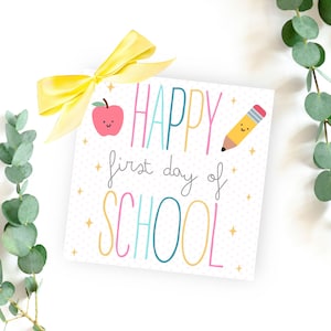 May include: A white card with the text "HAPPY first day of SCHOOL" in colorful letters. A red apple with a smiling face and a yellow pencil with a smiling face are on the card. The card is decorated with gold glitter.