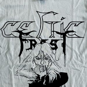 Op de afbeelding: Crèmekleurig T-shirt met een zwarte afbeelding van de band Celtic Frost. Het ontwerp bevat de bandnaam en een gezichtsillustratie. De tekst "PREPARE FOR THE BLITZ TOUR 1986" is eronder gedrukt.