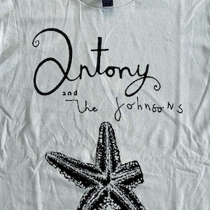 以下が含まれることがあります： オフホワイトのTシャツ。黒い筆記体で「Antony and the Johnsons」の文字が書かれ、その下に白黒のヒトデのイラストが描かれています。ヒトデの下には、手書き風の「Cripple and the Starfish」の文字が書かれています。