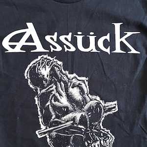 May include: A black short-sleeve t-shirt featuring a white graphic print. The top of the shirt displays the text "Assück" in a stylized font, while the bottom features the text "MISERY INDEX" with a central illustration of a seated figure.