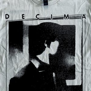 Puede incluir: Camiseta blanca con un gráfico en blanco y negro de una persona con traje, enmarcada en un rectángulo. Las palabras "DECIMA" y "VICTIMA" están impresas encima y debajo del gráfico.