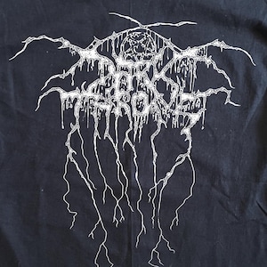 Może przedstawiać: Czarny t-shirt z krótkim rękawem z białym nadrukiem graficznym na klatce piersiowej. Wzór przedstawia napis Darkthrone stylizowaną, poszarpaną, ściekającą czcionką z liniami przypominającymi błyskawice rozciągającymi się w dół od liter.