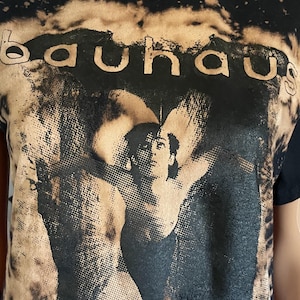 Op de afbeelding: Een zwart t-shirt met korte mouwen en een gebleekt, versleten effect langs de randen. Het midden toont een grafische print van een persoon met uitgestrekte armen, met daarboven de tekst "bauhaus" in een schreefloos lettertype.