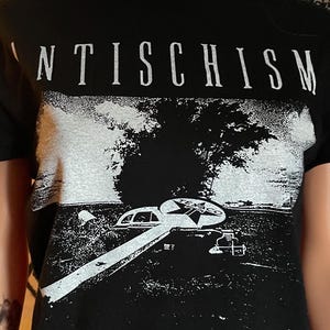 May include: A black short-sleeved t-shirt featuring the white text ANTISCHISM at the top. Below the text is a white graphic of a landscape with a tree and a circular emblem. Three identical circular symbols are printed in a row at the bottom.