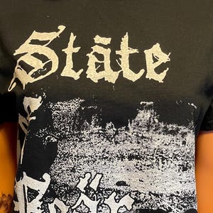 May include: A black short-sleeve t-shirt featuring a white graphic print. The text "State" appears at the top, with "Fear" below it, and "Wallow Squalor" printed at the bottom. The design includes a textured, distressed black and white image.