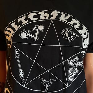 May include: A black short-sleeve t-shirt featuring a white graphic print of a pentagram inside a circle. The text "WITCHFINDER" is arched above the circle, and "GIVE 'EM HELL" is printed below. The pentagram contains small icons of axes, a goat head, a sword, and a winged serpent.