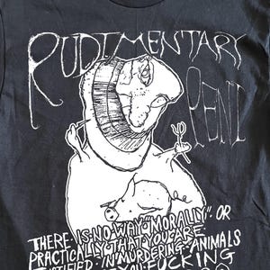 Op de afbeelding: Zwart t-shirt met witte grafische tekst en illustratie. De tekst luidt "RUDIMENTARY PENI" boven een tekening van een wezen. Daaronder staat: "THERE IS NO WAY MORALLY OR PRACTICALLY THAT YOU ARE JUSTIFIED IN MURDERING ANIMALS SO WHY DONT YOU FUCKING STOP ???"