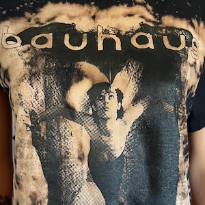 Peut inclure: Un t-shirt noir à manches courtes avec un effet délavé et usé autour du motif central. Le motif affiche le mot "bauhaus" en minuscules au-dessus d'une image en noir et blanc en demi-teintes représentant une personne aux bras écartés.