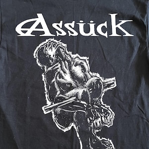 Puede incluir: Camiseta negra con gráfico y texto en blanco. El gráfico representa una figura sentada y angustiada que sostiene una barra. El texto "Assück" está encima del gráfico y "Misery Index" debajo.