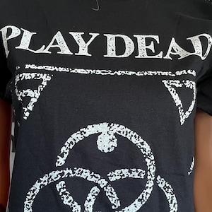 May include: A black short-sleeve t-shirt featuring the white text PLAY DEAD at the top. Below the text is a white graphic design consisting of three interlocking circles surrounded by a rectangular border with small triangles in each corner.
