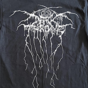 Puede incluir: Camiseta negra con un dise&ntilde;o gr&aacute;fico blanco. El dise&ntilde;o presenta texto estilizado y formas abstractas, con un pentagrama en la parte superior. La camiseta est&aacute; hecha de un material suave y tiene cuello redondo.