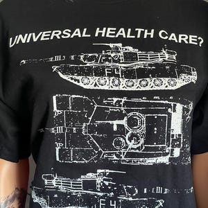 以下が含まれることがあります： 軍用戦車の白いスクリーン印刷グラフィックが施された黒の半袖Tシャツ。上部には「UNIVERSAL HEALTH CARE?」というテキストがプリントされており、グラフィックデザインの右下隅には「NO」という単語がプリントされています。