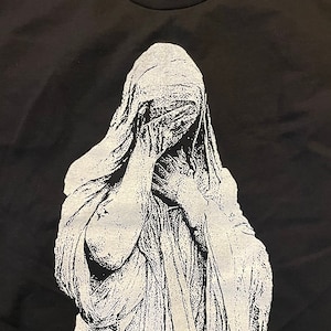 May include: Black t-shirt featuring a white graphic of a veiled figure covering their face with their hands. The design is a detailed, artistic illustration. The shirt has a crew neck and short sleeves.