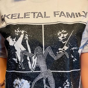 Może przedstawiać: Jasnoszary t-shirt z krótkim rękawem z czarnym nadrukiem graficznym. Napis "SKELETAL FAMILY" znajduje się u góry, a "SHE CRIES ALONE" u dołu. Centralny wzór składa się z czterech paneli przedstawiających członków zespołu oraz centralną postać.