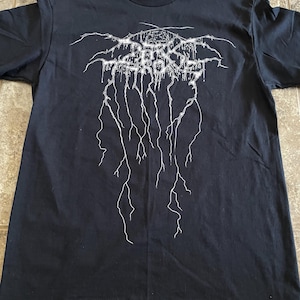 Puede incluir: Camiseta negra con un dise&ntilde;o gr&aacute;fico blanco. El dise&ntilde;o presenta texto estilizado y l&iacute;neas en forma de rayo. La camiseta est&aacute; hecha de un material suave y tiene cuello redondo.