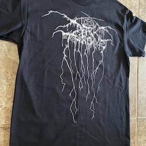 Puede incluir: Camiseta negra con un dise&ntilde;o gr&aacute;fico blanco. El dise&ntilde;o presenta el texto "DARK SORROW" en una fuente estilizada y goteante, con l&iacute;neas ramificadas que se extienden hacia abajo. La camiseta est&aacute; hecha de un material suave.