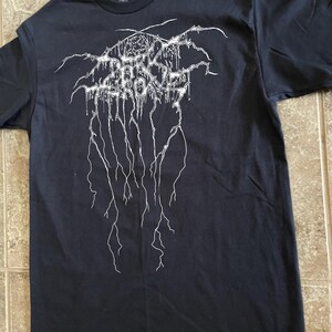 Puede incluir: Camiseta negra con un dise&ntilde;o gr&aacute;fico blanco. El dise&ntilde;o presenta texto estilizado y l&iacute;neas goteantes. La camiseta est&aacute; hecha de un material suave y tiene cuello redondo.