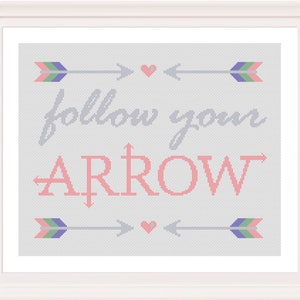 May include: A framed cross-stitch artwork with the words "follow your ARROW" in gray and pink. The design includes arrows with green, purple, and pink details, and small pink hearts. The artwork is set against a light gray background.