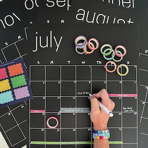 May include: Black chalkboard-style calendar with white text, featuring the month of July. Colorful washi tape and sticky notes are included. A person is writing on the calendar with a white marker. A colorful bracelet is visible.