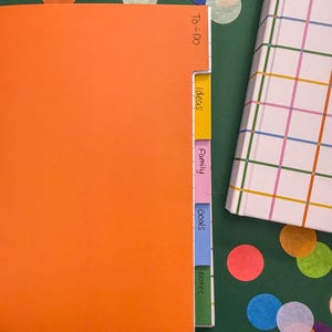 May include: An orange notebook with five colored tabs labeled To-Do, Ideas, Family, Goals, and Notes, positioned next to a white notebook with a multicolored grid pattern. The tabs are yellow, pink, blue, and green, with black handwritten text.