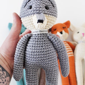 Pode incluir: Um brinquedo de pel&uacute;cia de lobo cinza crochetado com um rosto branco e marrom. O lobo est&aacute; segurando uma etiqueta que diz "Fert&eacute;-Bernard".
