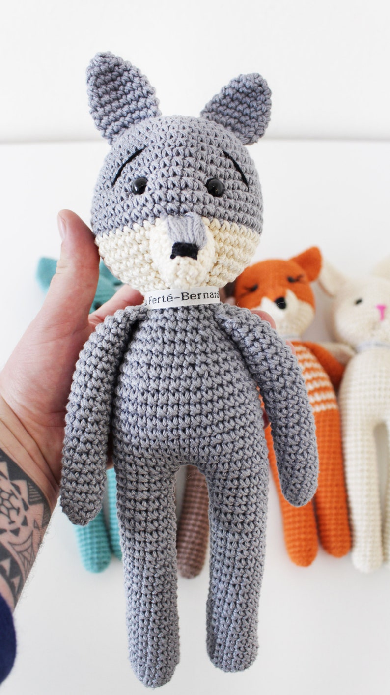 Pode incluir: Um brinquedo de pel&uacute;cia de lobo cinza crochetado com um rosto branco e marrom. O lobo est&aacute; segurando uma etiqueta que diz "Fert&eacute;-Bernard".
