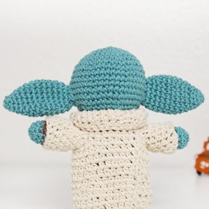Puede incluir: Un juguete de peluche tejido a crochet con forma de un personaje ficticio popular. El juguete lleva una bata de color crema y tiene orejas grandes y ca&iacute;das. El juguete est&aacute; hecho con hilo azul claro y marr&oacute;n.