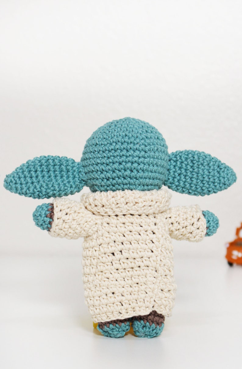 Puede incluir: Un juguete de peluche tejido a crochet con forma de un personaje ficticio popular. El juguete lleva una bata de color crema y tiene orejas grandes y ca&iacute;das. El juguete est&aacute; hecho con hilo azul claro y marr&oacute;n.