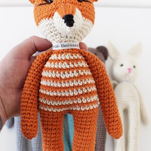 Pode incluir: Um brinquedo de raposa de croch&ecirc; laranja com listras brancas e olhos pretos. A raposa est&aacute; segurando uma etiqueta branca com o texto "Art&egrave;-Bernard".