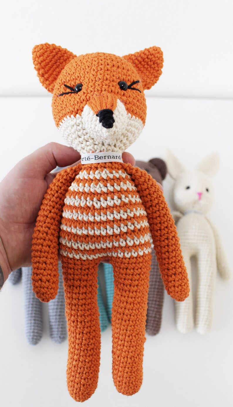 Pode incluir: Um brinquedo de raposa de croch&ecirc; laranja com listras brancas e olhos pretos. A raposa est&aacute; segurando uma etiqueta branca com o texto "Art&egrave;-Bernard".