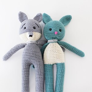 Pode incluir: Dois animais de peluche de croch&ecirc;, um lobo cinza e um coelho azul turquesa, de p&eacute; lado a lado. Ambos t&ecirc;m detalhes brancos e olhos pretos. O lobo tem um focinho branco e o coelho tem um nariz rosa.