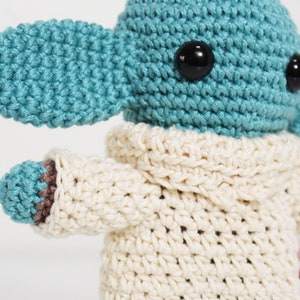 Puede incluir: Un juguete de peluche tejido a crochet con forma de personaje con orejas grandes, que lleva un su&eacute;ter tejido a crochet blanco. El juguete tiene un cuerpo azul y ojos negros.