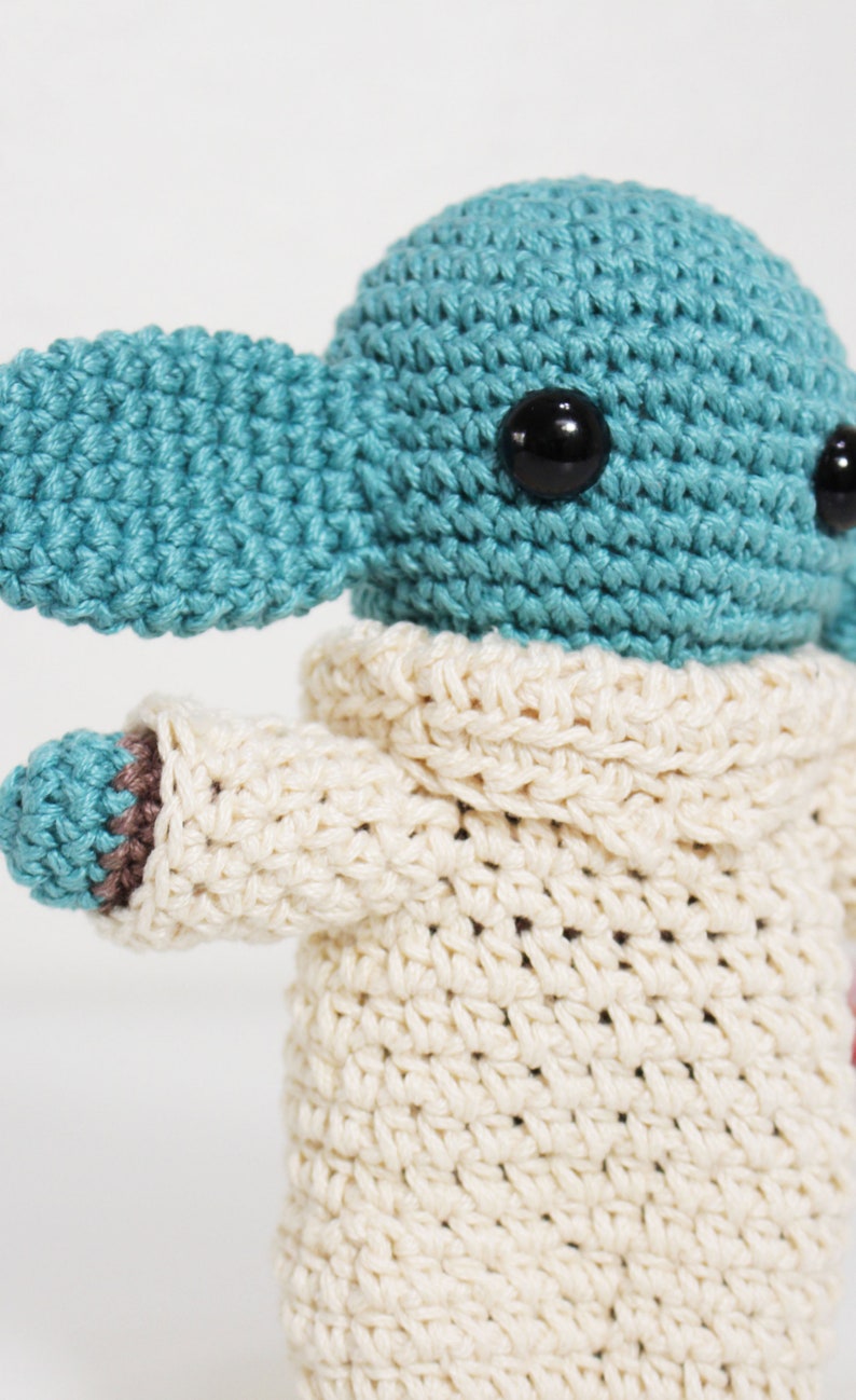 Puede incluir: Un juguete de peluche tejido a crochet con forma de personaje con orejas grandes, que lleva un su&eacute;ter tejido a crochet blanco. El juguete tiene un cuerpo azul y ojos negros.