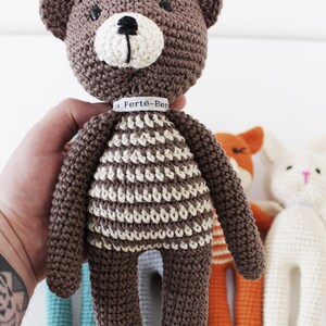 Pode incluir: Um urso de peluche de croch&ecirc; marrom com roupas listradas brancas e marrons. O urso tem olhos pretos e um nariz preto. O urso est&aacute; segurando uma etiqueta branca com o texto "La Fert&eacute;-Ben" nela.