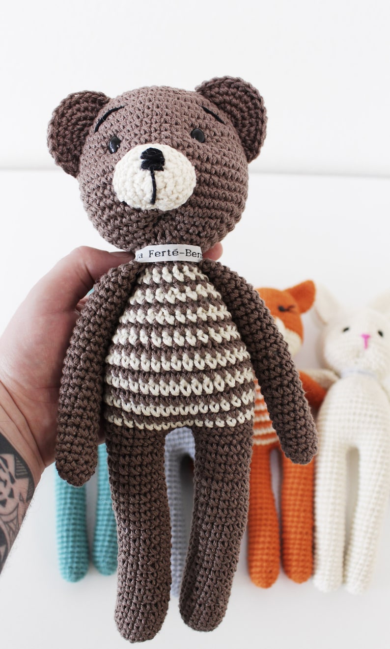 Pode incluir: Um urso de peluche de croch&ecirc; marrom com roupas listradas brancas e marrons. O urso tem olhos pretos e um nariz preto. O urso est&aacute; segurando uma etiqueta branca com o texto "La Fert&eacute;-Ben" nela.
