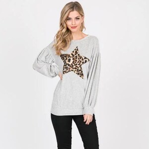 Puede incluir: Una sudadera gris con una gran estrella de estampado de leopardo en la parte delantera. La sudadera tiene mangas largas y un corte holgado.