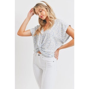 May include: A light blue short-sleeved blouse with a white floral print and a twist detail at the front. The blouse is worn with white jeans.