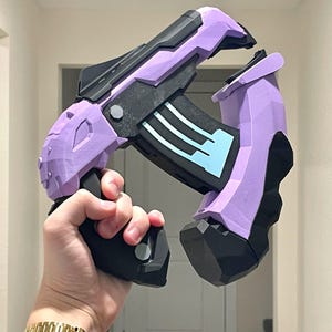May include: A futuristic-looking toy gun in shades of purple and black. The gun features a geometric design with angular shapes and a handle. The barrel has a black section with blue accents. The toy is held in a hand.