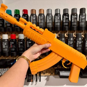May include: A bright orange toy rifle featuring a textured barrel, a curved magazine, a trigger guard, and a pistol grip. The object is constructed with a matte finish and displays a distinct geometric design with visible seams along the frame.