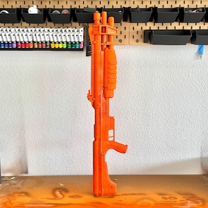 May include: Bright orange toy gun with a detailed design, standing upright on a wooden surface. The background includes a pegboard with storage containers, paint tubes, and spray paint cans. The gun's design features a handle, barrel, and various attachments.