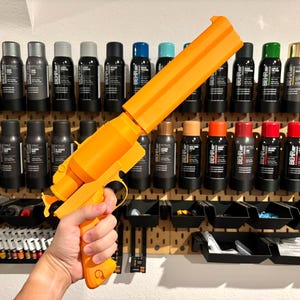 May include: An orange 3D-printed toy gun is held in front of a pegboard filled with paint bottles. The gun has a large barrel and a pistol grip. The paint bottles are black with white labels and various colored caps.