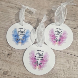 May include: Three white, circular ornaments with a ribbon for hanging. Each features a floral design and the text "I HAVE AN Angel IN HEAVEN I CALL HIM Grandad" or "Mum". The floral designs are blue, pink, and purple.