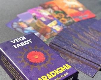 May include: A purple Vedi Tarot deck box titled Paradigma by Natalia Anani, featuring a red flower graphic. Several tarot cards with colorful illustrations are fanned out behind the box, displaying various figures and floral imagery.