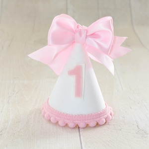 May include: A white party hat with a pink bow and the number 1. The base of the hat is trimmed with pink pom-poms. The hat is designed for a first birthday celebration.