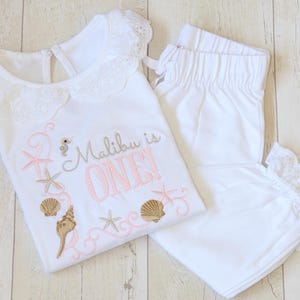May include: White baby outfit with a pink and brown embroidered design that says "Malibu is ONE!" with seashells and a seahorse. The outfit includes a top with a lace collar and matching trousers with lace trim.