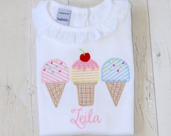 Puede incluir: Un body blanco para bebé con un cuello de volantes y tres conos de helado bordados. Los conos de helado son rosa, amarillo y azul con chispas y una cereza en la parte superior. El body está personalizado con el nombre "Leila" en rosa.
