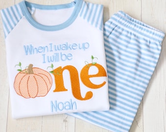 May include: A white pajama set with light blue striped sleeves and pants. The shirt features an embroidered orange pumpkin, the text "When I wake up I will be one" in light blue, and the name "Noah" embroidered in light blue below the pumpkin.