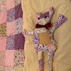 May include: A patchwork quilt with a floral pattern in shades of pink, purple, and white. A handmade patchwork toy in the shape of a bear is lying on top of the quilt. The bear is made from fabric with a floral pattern in shades of purple and white.